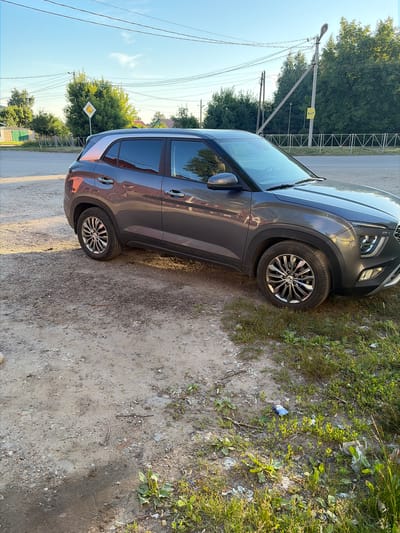 A grey Hyundai Creta SUV is parked on a dirt and gravel shoulder next to a paved road in Skopin, Russia. The car is positioned at a slight angle, facing towards the right of the frame, with its rear left wheel and front right wheel visible. The vehicle's doors are closed, and there are no people or animals in or around it.

The background shows a rural or suburban landscape. A white diamond-shaped road sign with a yellow border is visible in the distance, to the left of the car. Further back, there are trees and buildings, including a green shed or garage. Utility poles and power lines are also present, stretching across the sky. The ground in the foreground is a mixture of dirt, gravel, and sparse green vegetation. Some litter, including a blue packet and a white plastic bag, is scattered on the ground near the grass. The sky is clear and blue, with soft sunlight casting shadows.