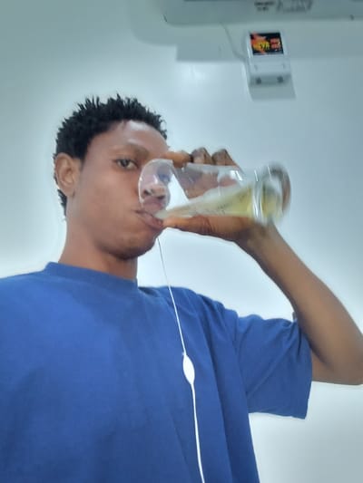 The image shows a young Black man in a blue t-shirt drinking from a glass. He is looking directly at the camera, with his left hand holding the glass to his mouth and his right hand supporting the bottom of the glass. There is a white wire cord hanging from his neck, possibly connected to headphones or a phone. He is in a room with white walls and a white ceiling. There is a white air conditioning unit mounted on the ceiling, and a white rectangular box is on the wall to the right of the camera. The coordinates provided place this image in Ujevwu, Nigeria.