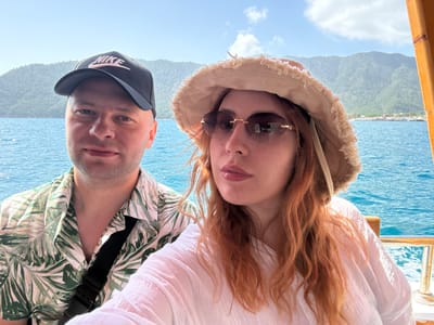 A couple is taking a selfie on a boat. The woman has long, red hair and is wearing a white shirt, a beige hat with a fringed brim, and sunglasses. The man is wearing a green and white leaf-patterned shirt and a black "Nike" baseball cap with a pink logo. They are positioned close together, facing the camera. The background shows blue water, a mountainous coastline, and a clear sky with some clouds. The location appears to be near Kumluca, Turkey, based on the landscape visible in the background.