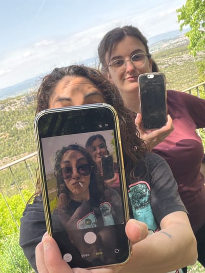 The image, oriented sideways, captures two individuals, likely young women, outdoors at a scenic overlook in Olèrdola, Spain.
The primary subject in the foreground is holding a smartphone, capturing a reflection. The phone's screen prominently displays a "selfieception," showing the foreground woman and a second person reflected, within a camera interface. The foreground woman has dark, curly hair, wears sunglasses, and has a lit cigarette in her mouth. She is wearing a dark t-shirt with a light blue and green graphic design. A small, unreadable tattoo is visible on her right forearm.
The second individual, partially visible behind the foreground woman and also reflected in the phone screen, has dark, straight hair and wears round-rimmed glasses. She appears to be holding a second smartphone, which is partially visible. This phone's screen displays "Favorites" and a "+" icon, suggesting an app menu.
The activity depicted is taking a reflection selfie, creating a recursive visual effect. The phone screen in the foreground shows various camera interface elements, including "0.5" and "x1" zoom options, a shutter button, and the words "VIDEO" and "FOTO" (Spanish for "photo").
The setting is an outdoor, elevated location, offering a wide view of a lush green landscape below, possibly hills or mountains covered in trees. The background shows a hazy distant horizon and bright natural light, indicating daytime. The scene suggests a relaxed moment enjoying a scenic view.