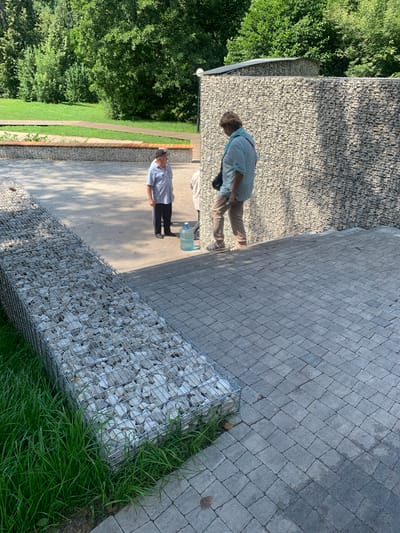 In Arbuzov-Baran, Russia, two men and one woman are present in an outdoor area with paved walkways and a gabion wall. The woman, wearing a light blue shirt and tan pants, is walking down a set of stone steps. To her left, a man in a checkered shirt and dark pants stands near a large water cooler. A second man, partially visible, is standing behind the woman. The gabion wall, filled with light-colored stones, forms a barrier to the right of the scene, and a similar gabion structure is visible on the left, filled with stones and bordering lush green grass. Trees and a grassy area are in the background.
