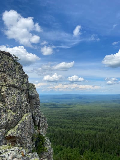 A high-angle, vertical medium shot captures a dramatic mountain landscape. In the foreground, a rough, gray rock face, partially covered in green lichen, dominates the left side of the frame. A small, sparse evergreen tree grows precariously from the top edge of the rock.
The middle ground reveals a vast, dense, dark green coniferous forest stretching to the horizon, with rolling hills and softer, muted green hues in the far distance. The sky above is a bright blue, adorned with scattered white cumulus clouds, some wispy cirrus streaks also visible. The image appears to be taken on a clear day, suggesting a natural, uninhabited environment. The location is identified as Mount Kruglaya in Magnitogorsk, Russia. There are no people or animals visible in the image.