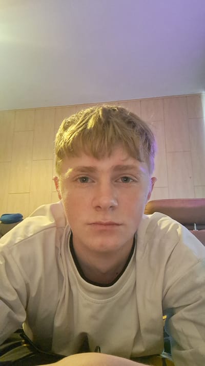 The media file is a close-up, self-portrait of a young person with light-colored hair and blue eyes. The subject is wearing a light-colored long-sleeved shirt. The background consists of light-colored tiled walls. To the right of the subject, a wall appears to be illuminated with purple light. The image is oriented vertically, with the subject's head to the right of the frame. The lighting suggests an indoor setting, possibly at night or in a dimly lit room. There is no visible text in the image. The subject's expression is neutral. The scene appears to be a casual, personal photograph taken by the subject themselves. The location context of Parchim, Germany, does not provide specific visual cues within the image itself.