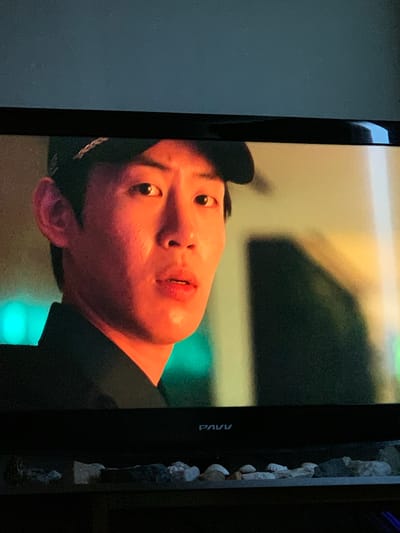 The image displays a close-up of a man's face on a television screen. The man appears to be East Asian with dark hair and is wearing a dark cap. He is looking directly at the camera with a slightly concerned or surprised expression. The lighting on his face is warm and orange-toned, highlighting his features. In the background, there are blurred elements of a room with some green and yellow lights. The television itself is mounted on a dark surface, and below the screen, a shelf with various rocks and shells is visible. The location of the television and its contents, as well as the broadcast being displayed, cannot be determined from the image provided.