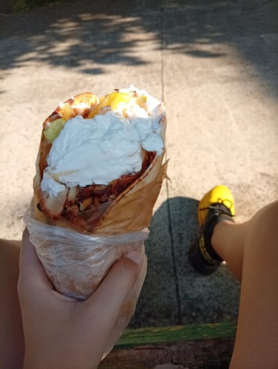 A person is holding a large wrap or gyros in their left hand, which is encased in a clear plastic bag. The wrap appears to contain meat, potatoes, lettuce, and a generous topping of white sauce, possibly yogurt or tzatziki. The person is sitting, as a leg is visible on the right, with a bright yellow shoe with black laces. The background shows a stone or concrete surface with dappled sunlight, suggesting an outdoor setting. The image appears to be taken in the city of Malko Chochoveni, Bulgaria.