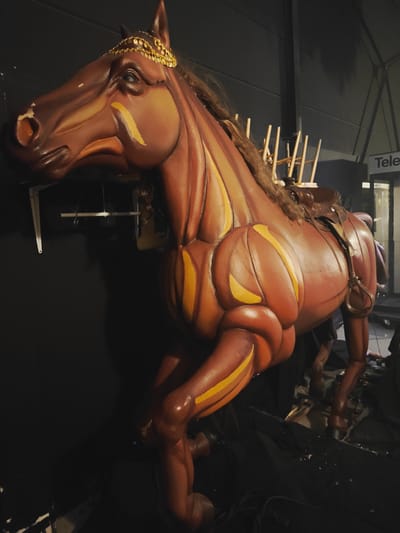 The image shows a large, brown, stylized horse puppet, likely a prop for a performance. The puppet is intricately detailed with sculpted muscles and is adorned with a gold headdress and a leather saddle. It is positioned in what appears to be a backstage area, with a dark, possibly black curtain behind it. There are some wooden support structures and wires visible around the puppet. In the background, a sign with the word "Tele" is partially visible, suggesting an indoor venue. The lighting is dim, casting shadows and highlighting the textures of the puppet. The overall impression is of a theatrical or performance-related setting in the city of Kirrwiller, France.