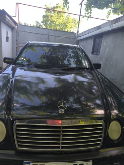 The image shows the front of a dark-colored Mercedes-Benz sedan parked in a somewhat cluttered outdoor area. The car's hood is dented and shows reflections of trees and foliage from above. A chrome grille with horizontal slats is prominent at the front, and a Mercedes-Benz star emblem is centered on the hood. The car has round, yellowed headlights. In the background, a grey, corrugated metal fence or wall runs horizontally, with a concrete structure and a small window visible to the left. To the right, a rough concrete wall with a white gutter runs along the top. Red piping runs across the top of the frame, possibly part of a structure or fence. Green leaves from trees overhang the scene. A license plate, partially visible at the bottom, displays Georgian characters and a small flag emblem. The location is identified as Pkhvenisi, Georgia. There are no people or animals visible in the image.