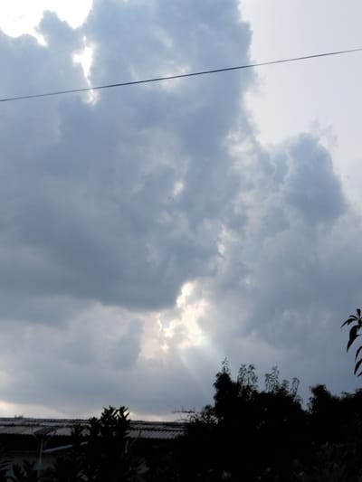 A dark, overcast sky dominates the frame, with the exception of a bright patch of light breaking through the clouds in the upper center. Sunbeams are visible radiating from this opening. A thin, dark horizontal line, likely a wire, stretches across the upper portion of the image. Silhouetted against the sky in the foreground are the dark shapes of trees and the top of a building with a corrugated roof. The overall impression is of a cloudy day with intermittent sunlight. No people are visible in the image. The scene is likely from an outdoor setting, possibly a residential area, given the presence of a building and trees. The location is identified as Nematjan, Uzbekistan.