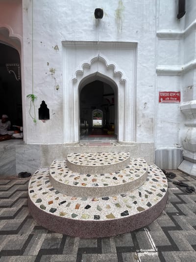The image captures a close-up view of the entrance to a religious building, possibly a mosque, located in Sylhet, Bangladesh. In the foreground, a three-tiered curved staircase with a terrazzo finish, embedded with colorful stones, leads up to the arched doorway. The steps are circular and the top two tiers are wider than the bottom one. The surrounding pavement features a dark gray zigzag pattern.
To the left of the entrance, a person wearing a white cap and traditional clothing is seated inside a dimly lit room, seemingly engaged in prayer or reading. A green string hangs from the wall near a small, dark niche. To the right of the entrance, a red sign with Bengali text and an arrow pointing upwards is affixed to the white wall. Several pairs of dark-colored sandals are placed on the pavement to the right and left of the staircase.
The building's exterior is painted white, showing some signs of wear and tear, including water stains and small marks. The entrance itself is an arched structure with decorative carvings. Beyond the immediate entrance, a long corridor or hallway is visible, leading deeper into the building. The overall atmosphere suggests a serene and spiritual setting.