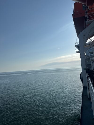 The image shows a view from the deck of a ship on a clear, sunny day. The sky is a bright, pale blue with some wispy white clouds low on the horizon. The sea is a dark, choppy blue-green, with gentle waves creating a textured surface. The edge of the ship is visible on the right side of the frame, with part of an orange life raft mounted high up. White railings and grey decking are also visible on the ship. The scene suggests a journey at sea, possibly a ferry or a larger vessel, with no land in sight. The location in the city of Unknown, Lithuania is not discernible from the image.