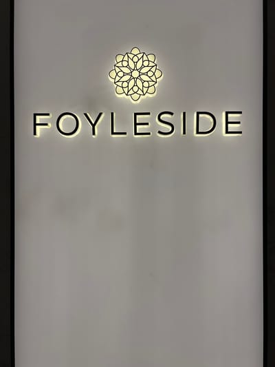 The image displays illuminated signage on a light-colored wall. Above the text "FOYLESIDE," there is a circular logo with a floral or mandala-like design, also illuminated. The "FOYLESIDE" text is in a sans-serif font with a black outline and a yellow-white glow emanating from behind the letters. The signage appears to be installed indoors, possibly in a mall or commercial building, given the clean, minimalist background. The city of Derry/Londonderry is known for the Foyleside Shopping Centre, a prominent retail hub. Therefore, this signage is likely located at the Foyleside Shopping Centre in Derry/Londonderry, United Kingdom. No people are visible in the image.