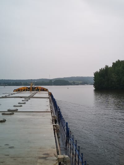 This image depicts a view from the deck of a cargo barge traveling on a wide river or canal. The barge's deck is mostly empty, made of concrete, with several closed hatches visible. A blue railing runs along the side of the barge. In the distance, a densely forested shoreline with rolling hills can be seen under an overcast sky. The water is calm with a slight ripple. The scene appears to be a tranquil, industrial waterway scene. The city of Goritsy, Russia, is a known location with waterways and industrial activity, making this a plausible setting for such a scene. There are no people visible in the image.