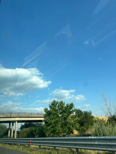 The image captures a view from inside a vehicle, looking out at a bright, sunny sky with scattered clouds. The sky dominates the left side of the frame, with faint reflections of the car's interior visible, including a circular object and diagonal lines. On the right side, a section of a highway guardrail runs vertically, with red reflectors visible. Beyond the guardrail, lush green trees and vegetation line the side of the road. In the background, a concrete bridge with metal railings spans across the landscape. The time of day appears to be late morning or early afternoon, given the bright sunlight and clear blue sky. The weather is fair and pleasant, suitable for travel. The scene suggests a journey along a highway in a rural or semi-rural area, possibly in Bulgaria, as indicated by the location context. There are no people or specific events depicted, but the image conveys a sense of movement and a peaceful, open-air environment.