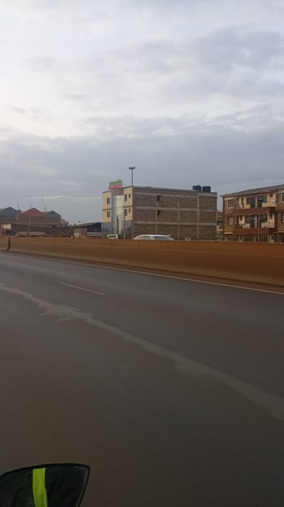A long, wet road stretches into the distance under a cloudy sky. A barrier runs alongside the road, obscuring the view of several buildings, including a multi-story structure with a sign that reads "New Meridian." Other structures include residential buildings. A portion of a vehicle with reflective stripes is visible in the lower-left corner of the image.