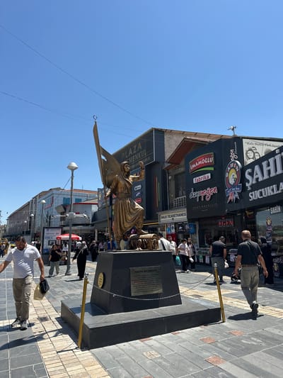 In Kayseri, Turkey, a bronze statue of a woman holding a flag and a sword stands prominently on a raised dark granite base in a pedestrianized urban area. The statue, depicting a historical or symbolic figure, is surrounded by shops and businesses with various signage in Turkish, including "PLAZA MENKUL," "İMAMOĞLU fabrika," and "ŞAHİN SUCUKLARI." The ground is paved with a patterned arrangement of grey and reddish-brown square tiles. Several individuals are visible walking along the street, some carrying bags. The sky above is clear blue. The statue is positioned in a public square or walkway, with shops and commercial establishments lining the sides of the area. The overall scene suggests a bustling city center on a bright, sunny day.