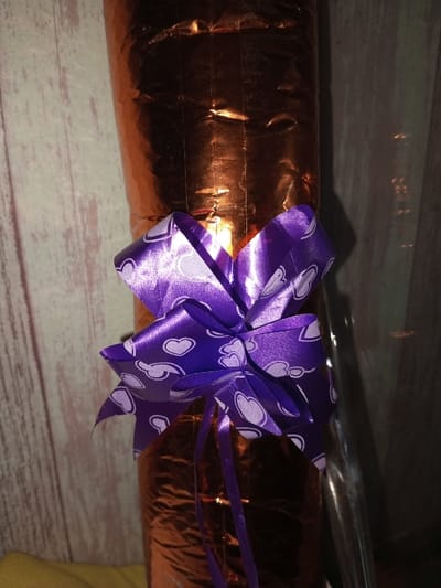 A gift, wrapped in orange metallic paper with visible creases and lines, is adorned with a bright purple satin ribbon. The ribbon is patterned with white hearts. The gift stands tall, with a light-colored wooden wall in the background. A hint of yellow fabric is visible at the bottom left. A shiny, clear material is on the right.