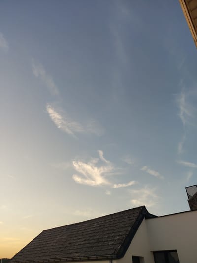 The image depicts a view of the sky from below, looking upwards. The sky is a pale blue, with scattered wisps of white clouds that appear thin and elongated. In the foreground, on the right side of the image, a dark, tiled roof of a building is visible, angled upwards and to the left. The building itself is white, with a dark-framed window visible on the upper section. On the left side of the image, a portion of another structure is visible, with a light-colored edge and a textured surface, also angled upwards. The lighting suggests it is either early morning or late afternoon, with the sun casting a warm glow towards the right side of the frame. There are no people or animals visible. The overall scene evokes a sense of quiet observation of the sky and surrounding architecture. The location is identified as Nederbrakel, Belgium, but there are no specific visual cues in the image to confirm this. No text is visible in the image.