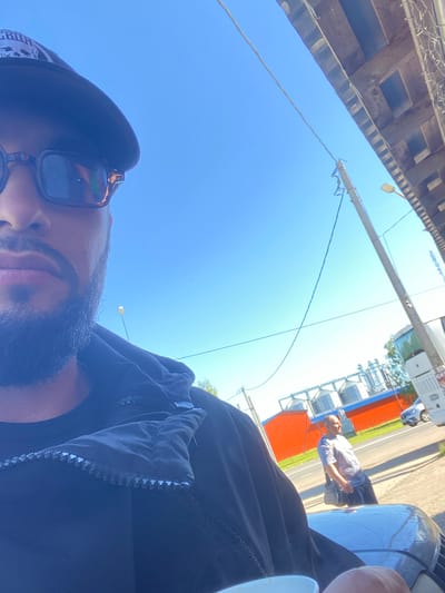 In Kaykino, Russia, a person in the foreground is taking a selfie outdoors during the daytime. The person is wearing a black baseball cap with a graphic design and dark-rimmed sunglasses. They have facial hair and are wearing a black jacket with a visible zipper. In the background, across a road, there are industrial silos with red and blue accents, and a person wearing a light-colored t-shirt and shorts is walking. A white bus and a silver car are also visible on the road. The sky is clear and blue. The scene appears to be along a roadside with some buildings or structures on the right side of the frame.
