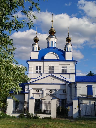 The image depicts a white and blue Orthodox church in Мамонтово, Russia. The church features three golden domes with spires, a blue roof, and white walls with blue trim. The building is surrounded by a black metal fence and some vegetation. A large leafy tree partially obscures the left side of the church. The sky is blue with scattered white clouds. The ground is covered with green grass.