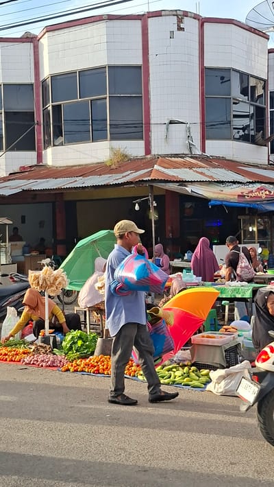 The image depicts an outdoor market scene in Keude Lueng Putu, Indonesia. In the foreground, a man wearing a blue shirt, gray pants, a beige cap, and black shoes walks from left to right, carrying two colorful woven bags. In front of him, laid out on the ground and tarps, are piles of fresh produce including tomatoes, onions, green beans, and cucumbers. A green umbrella is visible behind him.

In the mid-ground, several people are seen at market stalls. Some are wearing headscarves. One woman in a yellow shirt is seated behind a display of what appears to be snacks. Another woman in a pink hijab is standing near a stall. Further back, more individuals are present, with some interacting with vendors and others observing.

The background shows a building with a white tiled facade and red trim. The building has large, dark-paned windows and appears to be a commercial establishment. A corrugated metal roof covers the market stalls, suggesting a semi-permanent or temporary market structure. A motorcycle is partially visible on the far right of the image. The overall environment suggests a bustling local marketplace.
