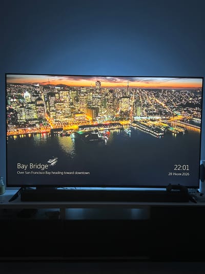 The image shows a television screen displaying a night-time aerial view of a cityscape, likely San Francisco, with the "Bay Bridge" labeled at the bottom left. The text "Over San Francisco Bay heading toward downtown" provides context. The time "22:01" and the date "28 Июля 2026" (July 28, 2026 in Russian) are also visible on the screen. The television is placed on a dark cabinet, with a soundbar positioned in front of it. The background is a dark blue wall, illuminated by a blue light from behind the television. The lower part of the image is dark, showing the cabinet and the floor, with a hint of a black game controller on the cabinet to the right. There are no people visible in the image. The city depicted is San Francisco, not Kapelle-op-den-Bos, Belgium.