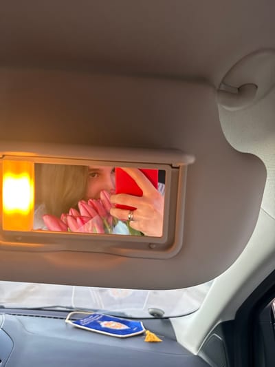 The image displays the interior of a vehicle, observed from a forward-looking perspective, presumed to be in the city of Slavyansk-na-Kubani, Russia. The primary subject is the driver's side sun visor, lowered to reveal an integrated vanity mirror with an illuminated orange-yellow light source on its left. Reflected in this mirror is a person, partially visible, holding a bouquet of pink tulips wrapped in clear plastic near their face. The person's right hand, adorned with a silver-colored ring on the ring finger, holds a vibrant red smartphone, angled as if capturing a selfie. The person's long, light brown hair is also visible in the reflection. Below the sun visor, a dark-toned dashboard is present. Resting on the dashboard is a blue fabric ornament, possibly a car charm, featuring an orange embroidered design and a yellow tassel. A small, dark, circular object is also discernible on the dashboard surface. The vehicle's ceiling and an overhead grab handle, both in a light beige or grey color, constitute the upper portion of the frame.