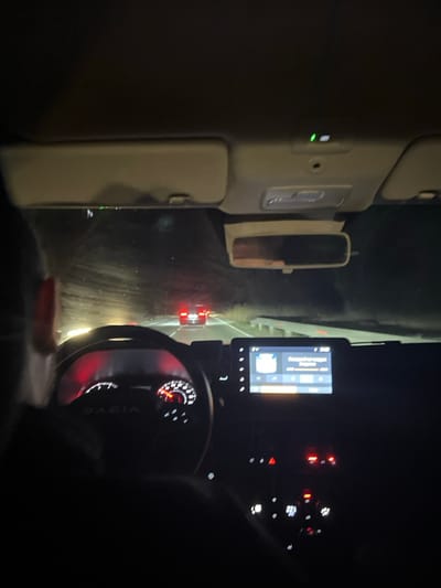 The image depicts the interior of a Dacia vehicle at night, viewed from the front passenger seat looking forward through the windshield. The scene is situated in Ushintsi, Bulgaria.

In the foreground and mid-ground, the driver's head, left shoulder, and upper arm are visible in silhouette on the left. The steering wheel, marked with the "DACIA" logo, is prominent, with its integrated instrument cluster displaying illuminated red and white gauges, including a speedometer and tachometer. To the right of the steering wheel, a central infotainment screen is visible, illuminated and displaying what appears to be a media player interface with text in Cyrillic script. Below the screen, multiple control buttons and dials are illuminated in red and white. Overhead, the vehicle's roof lining, sun visors, and a rearview mirror are visible.

Through the windshield, a dark, multi-lane road is illuminated by the vehicle's headlights. The red taillights of another vehicle, specifically an SUV, are clearly visible ahead in the center of the road. Further beyond, additional indistinct vehicle lights are discernible. A white guardrail runs along the right side of the road. The surrounding environment is dark, with hints of indistinct vegetation bordering the road, consistent with a nighttime rural or semi-rural setting. The windshield exhibits reflections and some streaking.