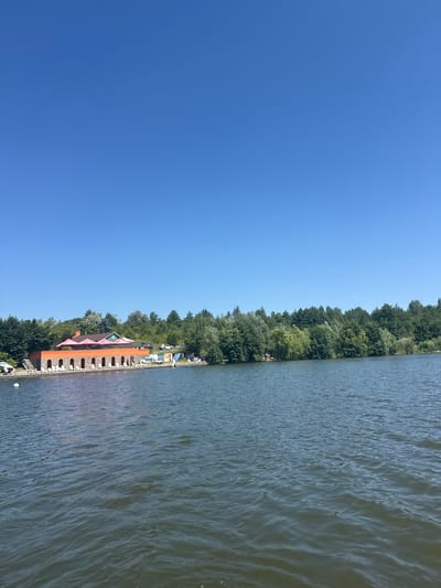 The image depicts a body of water with a resort-like establishment on the shore in Tiutky, Ukraine. The water occupies the lower half of the frame, showing gentle ripples and a dark, murky color. The shoreline features a long, orange-brick building with an arched facade, lined with white lounge chairs. Behind this building, a green-roofed structure is visible, and a line of lush green trees forms the backdrop. The sky above is a clear, deep blue. A few small boats are moored near the shore, and what appears to be a water slide is also present. The overall impression is a tranquil summer day at a lakeside resort.