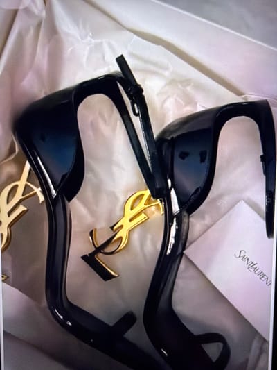 This is a high-angle close-up shot of a pair of black high-heeled Saint Laurent sandals placed inside a white box lined with white tissue paper. The sandals have a pointed toe and a thin ankle strap. Each sandal features a large, gold-colored YSL logo charm attached to the heel. The lighting is soft, creating a gentle glow and some reflections on the shiny surfaces of the sandals. A small, white card with the "SAINT LAURENT" logo printed in black script is partially visible on the right side of the frame, tucked beneath one of the shoes. The scene is devoid of any people or visible city elements, and therefore its location in Beșalma, Moldova, cannot be determined from the visual information provided.