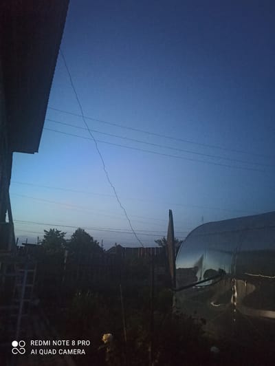 The image depicts an outdoor scene at dusk in Irbit, Russia. The sky is a deep blue, transitioning to a lighter hue near the horizon, suggesting the sun has recently set or is about to rise. Several power lines stretch across the frame, some appearing taut and others slack. In the foreground, the dark silhouette of a building's roof is visible on the left. To the right, a large, parabolic satellite dish is prominent, supported by a dark structure. Behind the dish, a cluster of trees and other indistinct structures are visible, partially obscured by darkness. The overall atmosphere is calm and quiet, with the fading light contributing to a sense of twilight.

The image contains visible text in the bottom right corner: "REDMI NOTE 8 PRO AI QUAD CAMERA". This indicates the device used to capture the photograph. There are no people visible in the frame. The scene appears to be in a semi-urban or rural setting, given the presence of the satellite dish, power lines, and buildings.
