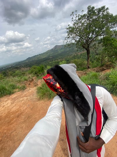 A person is reclining or resting on the reddish-brown earth of a hilltop, surrounded by patches of green grass and overlooking a vast natural landscape. The individual is dressed in a white long-sleeved top and white trousers. A gray blanket or garment with black faux fur trim covers part of their upper body. They wear a distinctive red patterned head covering, possibly a scarf or durag, featuring a pattern similar to the Louis Vuitton monogram. A dark-skinned hand rests on the gray garment.
In the midground, a prominent tree with lush green foliage stands, adding to the natural setting. The background reveals a panoramic view of distant hills or mountains blanketed in dense vegetation, stretching beneath an overcast but bright sky with scattered clouds. The scene suggests a daytime setting, possibly late morning or early afternoon. The overall atmosphere is one of peaceful relaxation, with the individual enjoying the expansive view from this high vantage point. The location is set in Amaigbo, Nigeria. No explicit readable text is visible in the image.