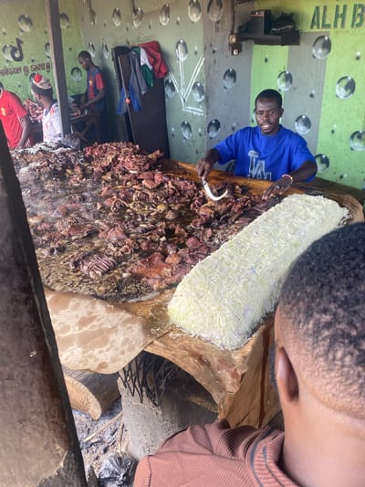 In Barakin Babele, Nigeria, a man in a blue t-shirt prepares a large quantity of fried meat on a wooden table. The meat, which appears to be stewed or boiled before frying, is spread out over a large surface and is steaming. Next to the meat is a mound of what looks like shredded cabbage or lettuce. The scene takes place in what appears to be an open-air food stall or market, with a green and white painted wall in the background decorated with circular patterns and lettering that partially reads "ALH B". Several other individuals are present, including a man in a red shirt and another in a black shirt and red shorts standing further back. The overall impression is one of a busy food preparation area in a Nigerian marketplace.