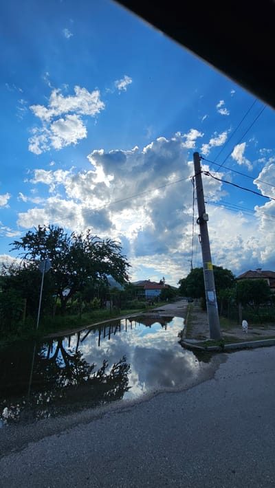 The image captures a vibrant, sunny day scene in Voiniagovo, Bulgaria, likely after rainfall, featuring a large puddle reflecting the sky. The perspective appears to be from a slightly elevated position, possibly from inside a vehicle, as a dark, angled structure partially obscures the top left corner.
The upper portion of the frame is dominated by a bright blue sky filled with scattered, fluffy white cumulus clouds. Sun rays are visible piercing through the clouds, especially towards the right side of the sky.
A tall, grey utility pole stands prominently in the mid-ground, slightly to the left of the center, with electrical wires extending across the frame. Two unreadable stickers, one yellow and one white, are affixed to the pole. Behind the pole and extending to the right, several houses with reddish-brown roofs and green vegetation are visible in the distance.
In the foreground and extending towards the mid-ground, a paved asphalt road stretches horizontally. A distinct concrete curb separates the road from a grassy, green verge to its left. A large, dark-leafed tree stands on the left side of the road, its branches reaching towards the sky.
The most striking feature is a significant puddle on the asphalt road in the lower right and center. This puddle brilliantly reflects the blue sky, white clouds, and distant trees and buildings, creating an inverted, mirror-like image of the overhead scene. In the distance, on the grassy verge near the road, a small white dog is visible. The overall impression is one of a peaceful, rural or semi-urban landscape on a bright day.