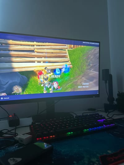 The media file displays a computer monitor showing a video game, likely Fortnite, given the visual style. The game screen shows a colorful, animated landscape with characters and building structures made of wood. In the center of the screen, there are two characters who appear to be ready for a game, indicated by the "READY" text on the screen. One character is wearing a yellow and blue outfit with a mask, while another character, a woman with red hair, is positioned above and to the right. The game interface also shows text indicating "SPECTATING: NATHANIEL123333A" and "FINDING NEXT MATCH".

To the right of the monitor, a gaming keyboard with illuminated keys, displaying a mix of purple, blue, and green lights, is visible. A gaming mousepad with a patterned design is also present beneath the keyboard. The overall setting appears to be a desk in a room, with the monitor and keyboard suggesting a gaming setup. There are also some electronic accessories, including a white power adapter, and a small black device with a digital display showing "88", which could be part of the computer setup. The lighting in the room appears to be dim, contributing to the visual prominence of the illuminated keyboard and monitor screen. The media file is oriented vertically, as if the photo was taken with a phone held in portrait mode.