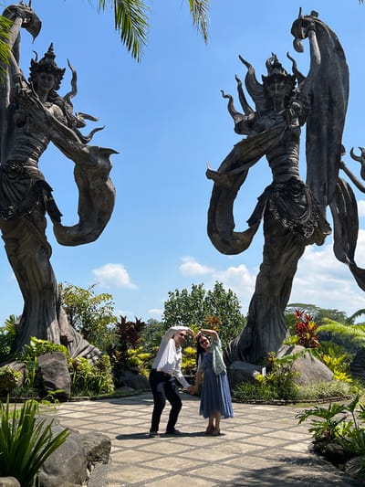 A couple is posing on a stone-tiled path in Kedewatan, Indonesia. The woman, on the right, wears a blue dress and a light-colored wrap. The man, to her left, wears a white shirt and dark pants. They are holding hands and forming a heart shape with their arms above their heads.

Two large, weathered stone sculptures flank the couple. The sculptures depict ornate, mythical figures with flowing drapery. Lush greenery, including trees, shrubs, and rocks, surrounds the path and sculptures. The sky is blue with scattered clouds.
