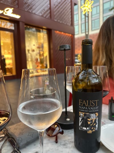 A bottle of Faust Napa Valley Cabernet Sauvignon 2022 rests horizontally on a wine rack alongside a stemmed glass, its surface beaded with condensation. The rack has a dark finish, contrasting with a light-colored marble-like surface. Sunglasses and eyeglasses are nearby. The background features a partially visible building exterior with a decorative, gold-toned starburst hanging from a light. The building has large windows displaying interior items.