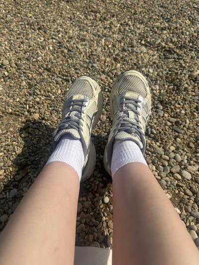 The image, taken from a top-down perspective, shows the lower legs and feet of a person standing on a surface covered with small, light-colored pebbles or gravel. Both legs are bare with fair skin, extending into white, ribbed ankle socks. The person is wearing light grey and white athletic shoes, which feature mesh uppers, synthetic overlays, and grey shoelaces. A subtle "ASICS" logo is visible on the side of both shoes, near the laces. Bright sunlight casts distinct shadows of the legs and feet onto the gravel, indicating a clear, sunny daytime environment. The setting appears to be outdoors, likely a path or trail within the Cukarica Urban Municipality in Serbia.