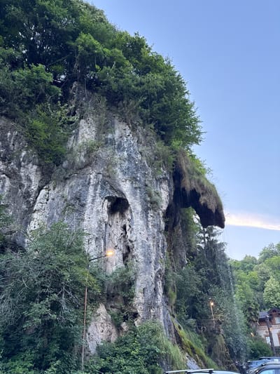 The image depicts a scenic natural landscape in Induno Olona, Italy, featuring a dramatic rock face with a waterfall cascading down its side. The rock formation is covered in lush green vegetation, with trees and moss growing in abundance. The waterfall itself is a prominent feature, with water flowing over the edge and creating a misty spray. In the foreground, a street light illuminates the area, and a few cars are parked below, suggesting this is a public access point or viewpoint. The sky above is a clear blue, with a faint wispy cloud. The overall impression is one of natural beauty and tranquility, with the waterfall adding a dynamic element to the scene.