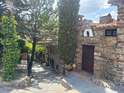 The image depicts a view of a narrow, stone-paved street in Patones De Arriba, Spain. On the right, a rustic stone building with a dark wooden door and a small, black-paned window is prominent. The building features a red-tiled roof and a decorative, abstract stone sculpture above the window. Green foliage and trees are visible throughout the scene, with ivy climbing a lamppost on the left and a tall cypress tree standing near the building. The street is partially shaded by the trees, creating a dappled light effect on the stone paving. In the distance, more stone buildings and vegetation suggest a traditional village setting. The sky is partly cloudy.