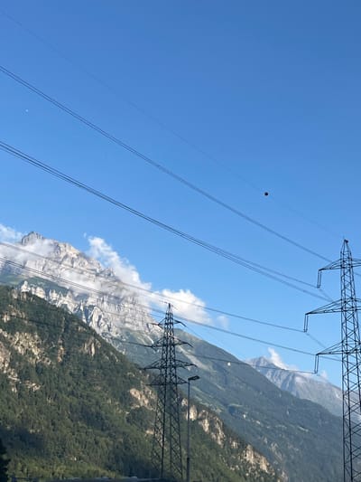 The image captures a panoramic view of a mountainous landscape in Erstfeld, Switzerland, under a clear, bright blue sky. Dominating the foreground and mid-ground are numerous parallel high-tension power lines, supported by two large, dark metal lattice transmission towers. A single, dark red spherical power line marker is visible on one of the higher tension lines in the upper right portion of the frame. In the upper left sky, a faint, pale crescent moon is discernible. The background features steep, rugged mountains, their lower slopes covered in dense green coniferous forest, transitioning to barren, grey, rocky peaks at higher elevations, some partially enveloped by white clouds. The scene is devoid of human figures or discernible activity.