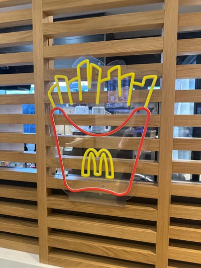 The image shows a close-up of a McDonald's sign made of neon lights, shaped like french fries and the Golden Arches logo. The sign is mounted on a wooden slat wall. The lighting is bright, and the sign is illuminated. The background appears to be an interior setting, possibly a McDonald's restaurant. The location is Saalfeld/Saale, Germany.