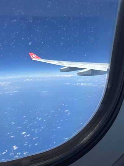 The image shows an airplane wing, viewed from inside the aircraft through a window. The wing has a red tip with a white emblem on it. The window frame is visible. Outside the window, there is a blue sky with some cloud cover. The picture appears to be taken mid-flight. The location, Tambon Sa Khu, Thailand, cannot be verified from the image.