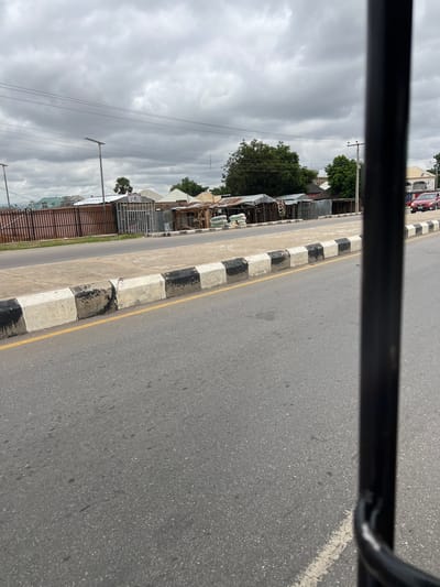 The image shows a street scene in Kporva, Nigeria. The foreground is dominated by a gray asphalt road with a yellow line and a concrete curb painted with alternating black and white blocks. In the middle ground, there is a sidewalk lined with a fence and various small roadside shops constructed from corrugated metal. Bags of what appear to be cement are stacked in front of some of the shops. The background features trees, buildings, power lines, and a cloudy sky. A red car is visible on the road to the right. The perspective suggests the photo was taken from inside a vehicle.