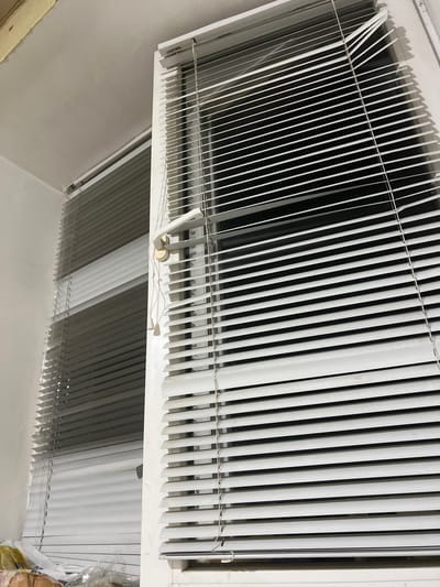 The image shows an interior view of a room with white blinds covering windows. The blinds are partially open, allowing glimpses of the dark window panes behind them. One set of blinds on the right is fully visible, with a white handle and pull cords hanging down. To the left, another set of blinds is visible, appearing to be a darker shade of gray. The ceiling is white and shows some signs of wear and tear. The scene appears to be a domestic setting, possibly a bedroom or living room. There are no people visible in the image. The location is identified as Luhansk, Ukraine.