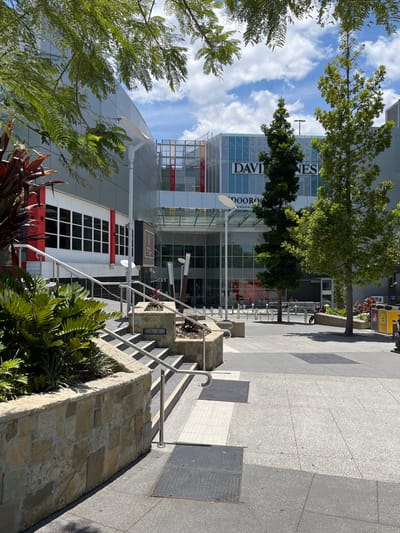 This image captures a vibrant, modern urban scene, likely a shopping precinct, under clear daylight. The central focus is a large, contemporary building featuring expansive glass facades and white and grey paneling, with hints of red accents. Prominently displayed on this building is the clear white text "DAVID JONES", indicating a department store. Below this, partial text reads "INDOOROOPILLY SHOPPING", confirming the location as Indooroopilly Shopping Centre in Australia.

In the foreground, a series of sturdy, stone-clad planters filled with lush green foliage, including ferns and broad-leafed plants, line the base of the image. A set of concrete stairs with sleek stainless steel handrails ascends from the lower right towards the center, leading up to a wide pedestrian pathway. This pathway, composed of light and dark grey pavers, extends into the distance, bordered by green trees that cast distinct shadows, suggesting a bright, sunny day, likely mid-morning or early afternoon. Manhole covers are visible on the paved ground.

Further into the scene, a light pole or signpost with a small white dish is present, and what appear to be bicycle racks can be glimpsed near the trees along the pathway. Above the buildings and trees, a bright blue sky is adorned with scattered, fluffy white clouds. The overall impression is one of a clean, well-maintained commercial area, devoid of visible people, conveying a quiet and serene atmosphere.