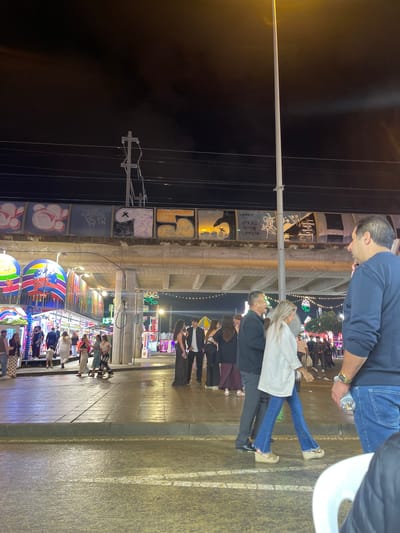 The image captures a nighttime scene in Jerez De La Frontera, Spain. A pole extends vertically from the lower left, reaching the top center of the image. The background reveals a bustling street, with people walking and standing in front of a brightly lit fairground or carnival. Buildings with graffiti are visible on the left side of the frame. A group of people, including a man in a blue shirt and jeans holding a water bottle, are positioned at the top of the image, partially obstructing the view of the fairground. Additional people are seen in the middle ground, standing on a paved surface.