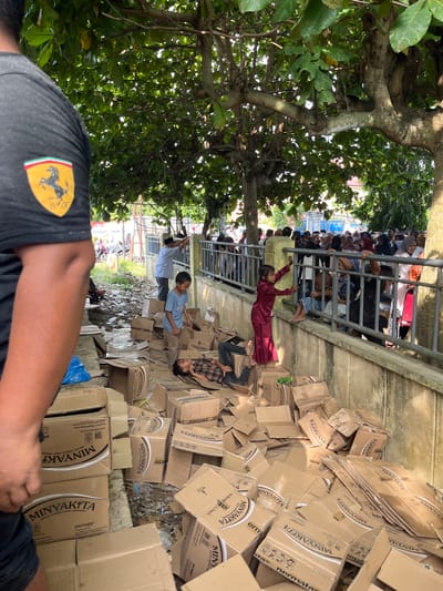 The image depicts a scene of disarray and possible distribution in Pulodolang, Indonesia. Numerous cardboard boxes, many labeled "MINYAKITA" and "permata", are scattered across the ground. Some boxes appear to be opened or damaged, with contents potentially spilling out.
In the foreground, the arm and shoulder of a person wearing a black t-shirt with a Ferrari logo is visible. Further into the scene, a young boy in a blue t-shirt and brown pants stands near a pile of boxes, looking towards the side. Another child, lying on their side on the cardboard, is visible in the lower center of the image.
Behind the boxes, a concrete wall separates the foreground from an area where a crowd of people are gathered, many of them women and children. They are leaning on a metal railing, suggesting they are waiting or observing. A young girl in a red outfit stands near the railing, reaching out. In the background, a few motorbikes are parked on a patch of grass, and the presence of trees suggests an outdoor setting. The ground is littered with debris and discarded materials, contributing to the overall sense of disorganization.