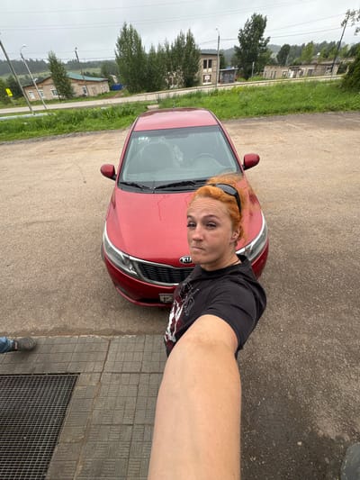 A woman with ginger hair and sunglasses on her head takes a selfie in front of a red Kia Rio. She is wearing a black t-shirt with a graphic print. The car is parked on a paved surface, likely a street or parking lot. In the background, there are buildings, trees, and a cloudy sky. The location is specified as Voznesenskoye, Russia. The overall scene suggests an outdoor setting, possibly in a town or village, with a focus on the woman and her vehicle.