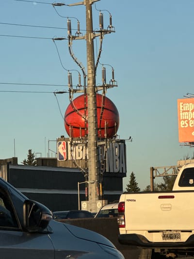 The image is taken outdoors in what appears to be Martínez, Argentina, based on the license plate "BGI-039" from Argentina visible on the back of a white Toyota pickup truck. The foreground contains the side of a dark-colored car. Prominently featured is an NBA-themed installation consisting of a large, red basketball-shaped structure and the word "NBA" in large block letters. The installation includes the NBA logo. An electrical pole with wires runs vertically in the image, seemingly bisecting the installation. An orange sign with text in Spanish ("Empe... es el") is visible above the NBA structure. The sky is a clear blue.