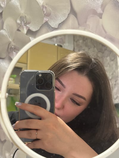 A young woman is taking a selfie in a mirror. She is holding a smartphone with a clear case in her left hand, angled to capture her reflection. She is wearing a thin ring on her left ring finger. Her right hand is not visible in the frame. The woman has dark brown hair and is wearing makeup, including eyeliner and well-defined eyebrows. The background of the selfie shows a wall with a floral wallpaper pattern featuring large, pale orchid-like flowers. In the reflection, the woman's face is partially obscured by the phone, and a blurred yellow wall is visible behind her. The mirror is round and has a white frame. The image appears to be taken indoors, possibly in a bathroom or bedroom. No specific location in Osentsy, Russia, can be identified from the image.
